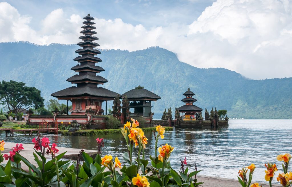 the view of a sacred temple in Bali called Ulun Danu Temple during the day with colorful flowers and a calm lake is perfect if you visit this place if doing outdoor activities in Bali