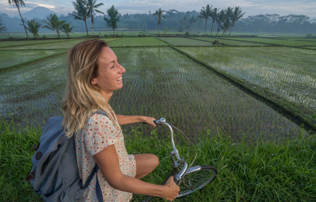 a woman wearing a white shirt with red and orange polka dot patterns is smiling happily while doing one of the outdoor activities in Bali, cycling with views of green rice fields