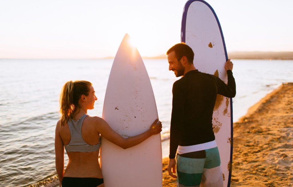 a man wearing a black long sleeve surf shirt and Tosca color pants holding a white surfboard and a woman wearing a gray and black bikini holding a white surfboard on a beach with an atmosphere before sunset doing one of the outdoor activities in Bali