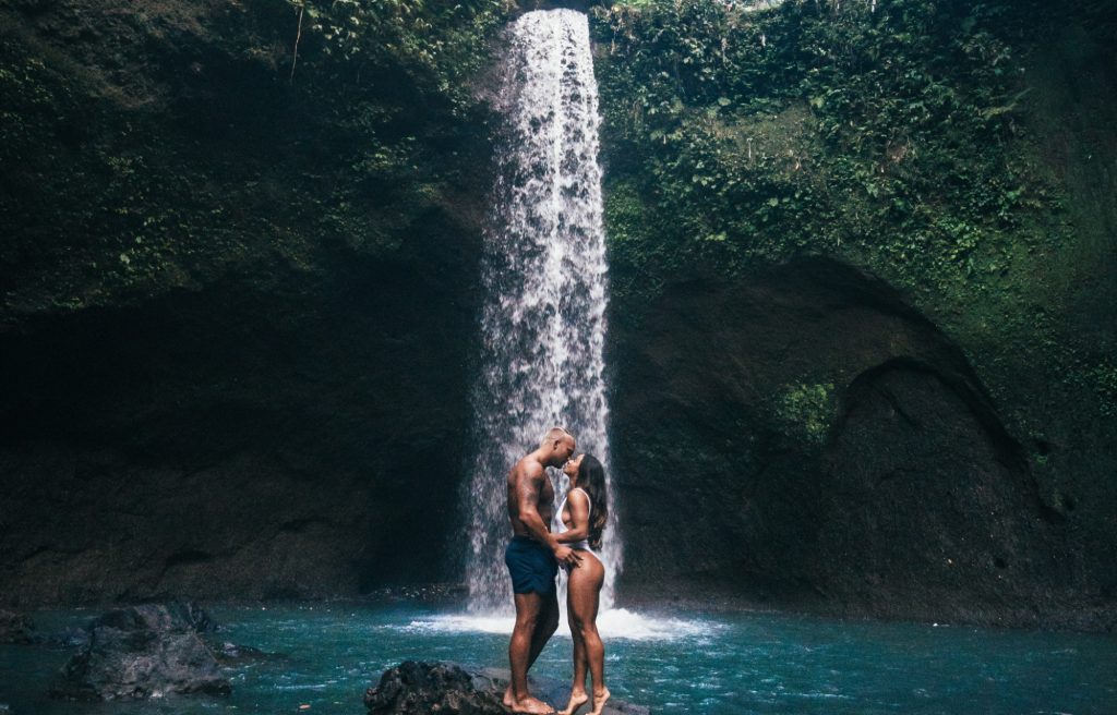 a man in blue shorts and a woman in a white bikini kiss in front of a waterfall during outdoor activities in Bali